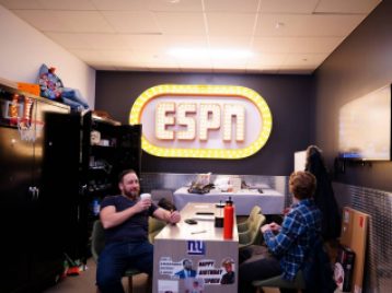 The ESPN logo on the back wall of a room, with two ESPN employees siting at a table having a coffee break.